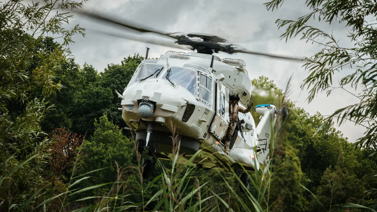 Een helikopter met openstaande deuren en een zwaaiende militair bij de uitgang vliegt laag door de bossen heen. 