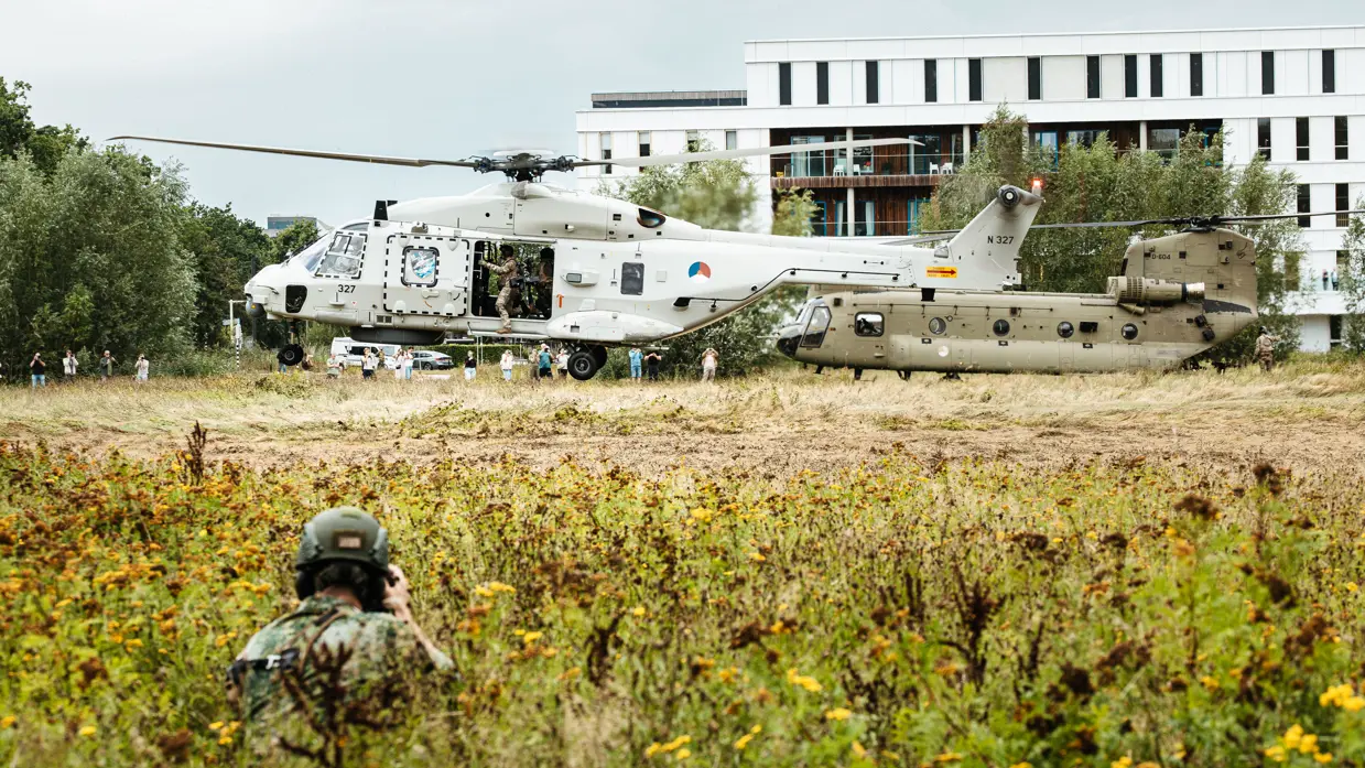 2 helikopters landen achter elkaar op een weide voor een ziekenhuis, gadegeslagen door een gehurkte en gecamoufleerde militair en aanwezige toeschouwers.