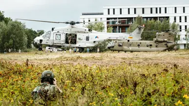 2 helikopters landen achter elkaar op een weide voor een ziekenhuis, gadegeslagen door een gehurkte en gecamoufleerde militair en aanwezige toeschouwers.