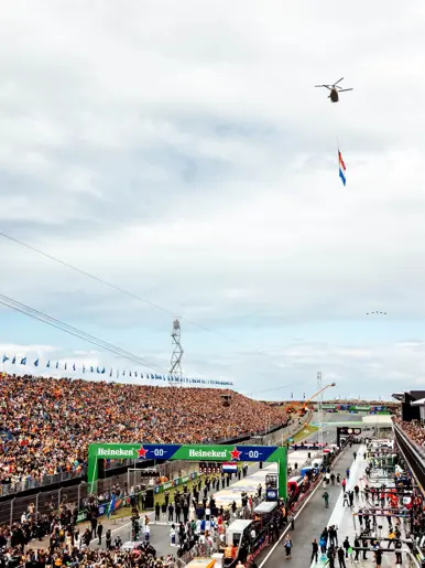 Circuit van Zandvoort met veel mensen op de baan en een grote gevulde tribune ernaast. Boven de baan vliegt een Chinook met daaronder een wapperende Nederlandse vlag.