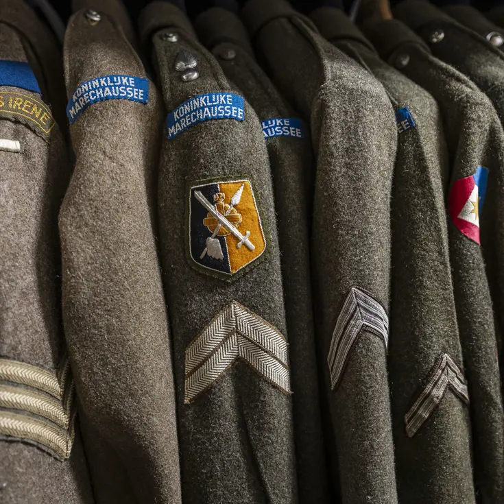 Close-up van een rij groene, wollen uniformen met een badge van de Koninklijke Marechaussee.
