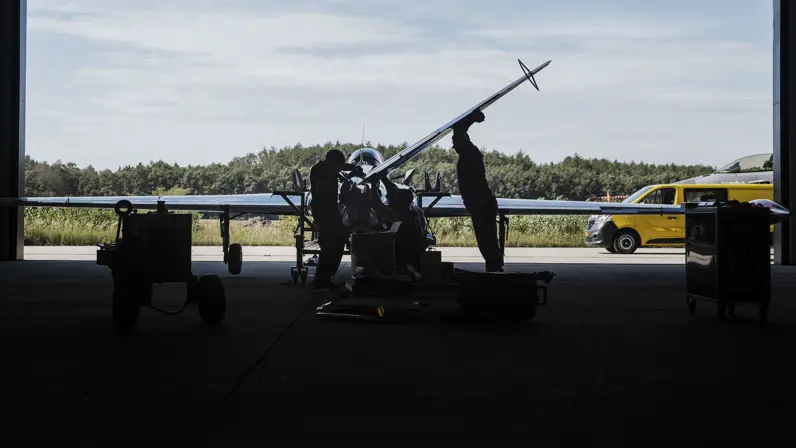 Silhouet van twee militairen die sleutelen aan een vliegtuig. Buiten de hangaar staat een geel busje.