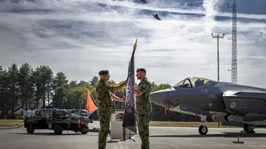 Windhouwer overhandigt aan Kerkhofs een vlag. Op de achtergrond de neus van een F-35 en een aanhanger met daarop F-35 munitie. Er vliegen twee F-35’s over.