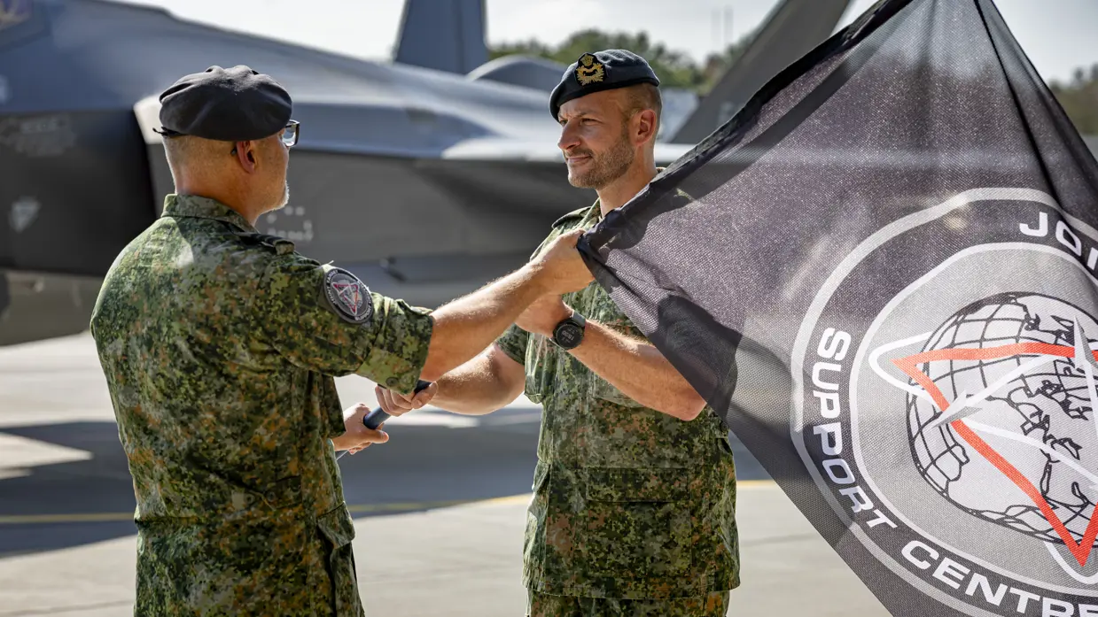 Kerkhofs neemt een vlag van Windhouwer in ontvangst. De vlag met daarop het embleem en de tekst ‘Joint Target Support Centre’ is daarbij half in beeld. Op de achtergrond staat een F-35.