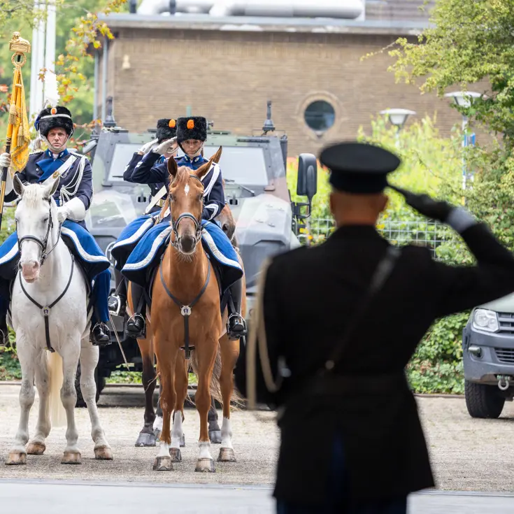 C-KMar Costongs van op de rug bezien, salueert naar ruiters te paard die teruggroeten.