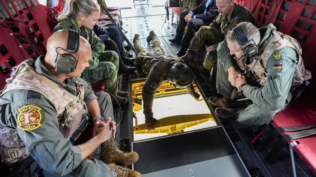 Steur en Neumann kijken door een open luik in de vloer van een Chinook vanaf een helikopterstoeltje aan de zijkant. Naast hen zitten nog twee militairen en drie burgers. Een loadmaster ligt languit op de vloer van de Chinook en kijkt door de opening.