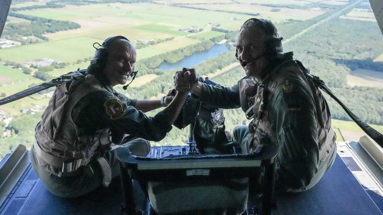 Steur en Neumann drukken elkaar de hand omkijkend terwijl ze op de klep van een vliegende Chinook zitten. 
