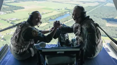 Steur en Neumann drukken elkaar de hand omkijkend terwijl ze op de klep van een vliegende Chinook zitten. 