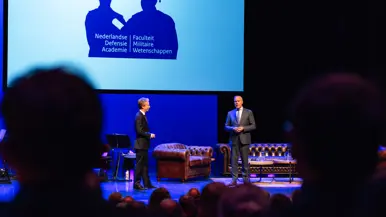 Twee mannen in burger pratend op een podium met achter hen een zithoek. Boven hen een groot scherm met daarop de tekst ‘Nederlandse Defensie Academie, Faculteit Militaire Wetenschappen’.