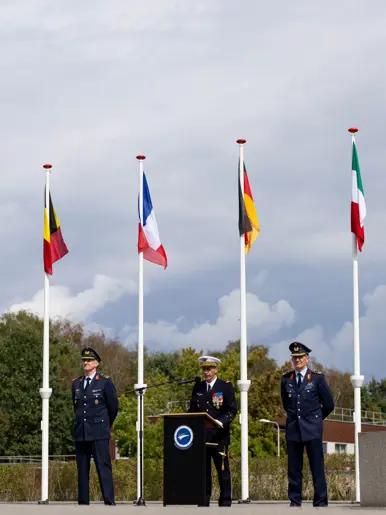 Drie militairen naast elkaar, waarvan de middelste spreekt vanachter een spreekgestoelte. Ze staan tussen vier grote vlaggenmasten met vlaggen van diverse EU-landen.