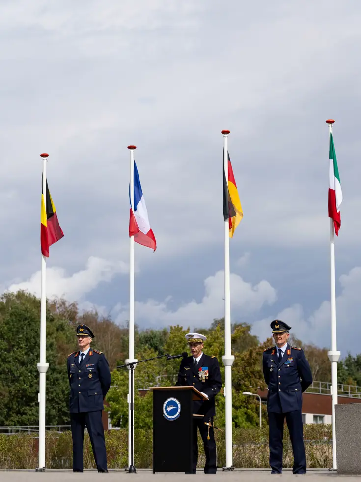 Drie militairen naast elkaar, waarvan de middelste spreekt vanachter een spreekgestoelte. Ze staan tussen vier grote vlaggenmasten met vlaggen van diverse EU-landen.