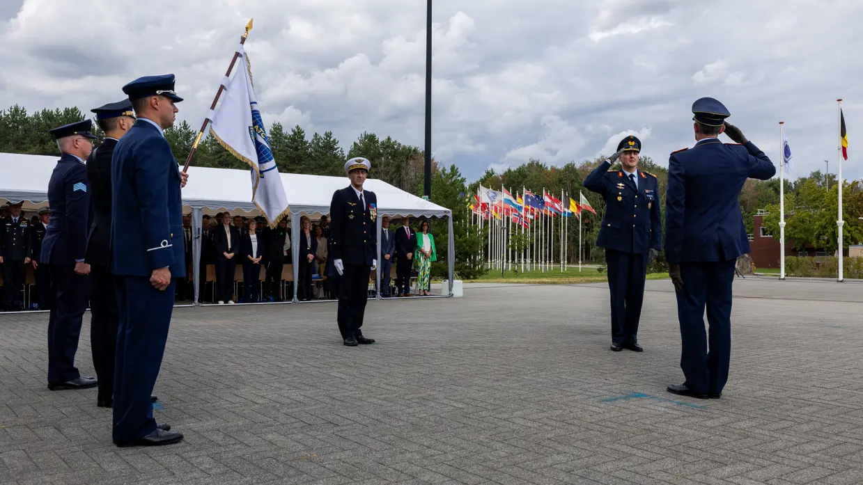 Drie militairen op een parade, twee van hen brengen elkaar de eregroet. Drie militairen kijken toe, de middelste draagt een vlag met het embleem van het EATC. Op de achtergrond staat publiek onder een lange witte partytent.
