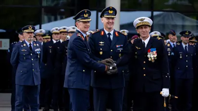 Drie militairen houden de handen opéén en kijken lachend op. Op de achtergrond staan meerdere rijen militairen opgesteld.