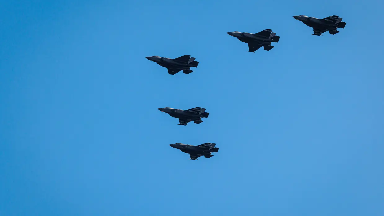 Vijf F-35’s in V-formatie de lucht tegen een strak heldere hemel. 