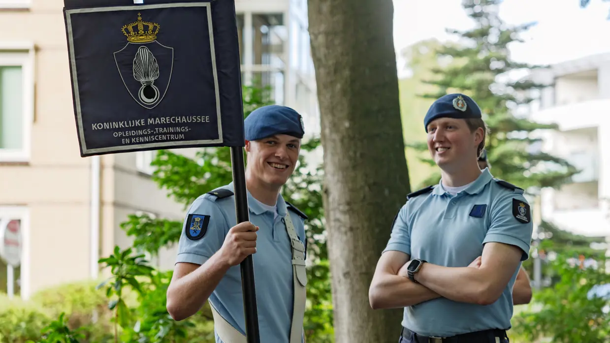 Twee marechaussees lachen naar de camera. De linker heeft een vaandel in de hand.