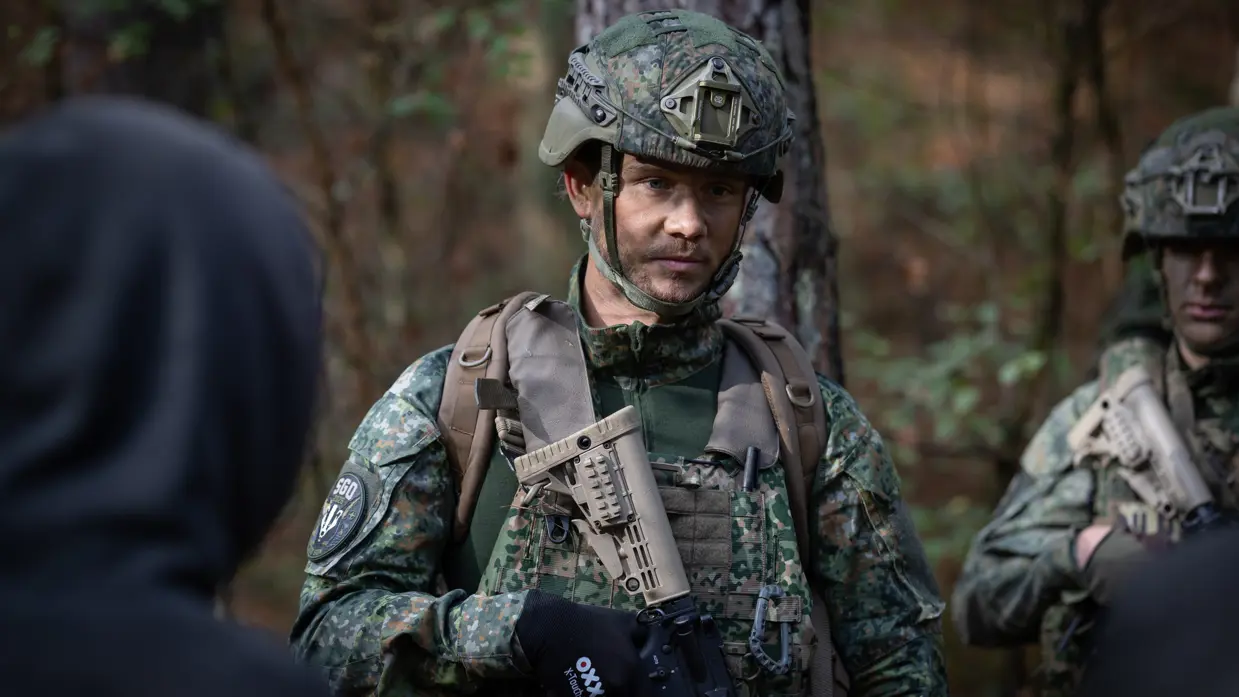 Een militair met stoppelbaard kijkt serieus voor zich uit. In zijn handen een wapen. Een blonde, vrouwelijke militair kijkt opzij en wijst.