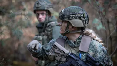 Een militair met stoppelbaard kijkt serieus voor zich uit. In zijn handen een wapen. Een blonde, vrouwelijke militair kijkt opzij en wijst.