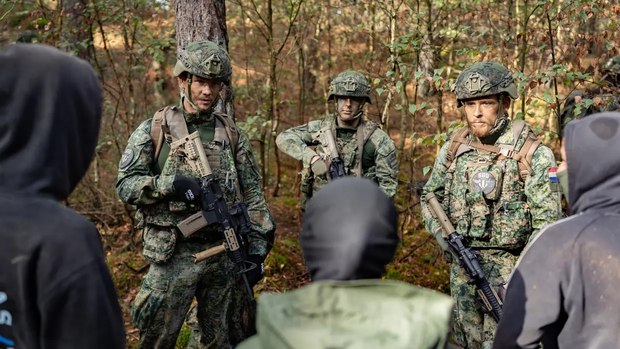 Drie gewapende militairen in gesprek met drie personen in donkere burgerkleding. Een vrouw met een donkere trui en een bedekt gezicht. Militairen in gevecht met de donker geklede partizanen. Twee militairen houden een persoon in burgerkleding vast terwijl diegene met zijn buik op de grond ligt.