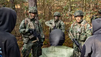 Drie gewapende militairen in gesprek met drie personen in donkere burgerkleding. Een vrouw met een donkere trui en een bedekt gezicht. Militairen in gevecht met de donker geklede partizanen. Twee militairen houden een persoon in burgerkleding vast terwijl diegene met zijn buik op de grond ligt.