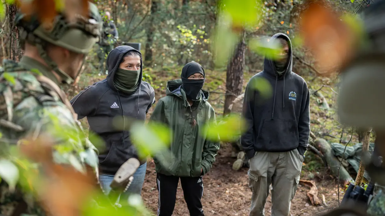 Drie gewapende militairen in gesprek met drie personen in donkere burgerkleding. Een vrouw met een donkere trui en een bedekt gezicht. Militairen in gevecht met de donker geklede partizanen. Twee militairen houden een persoon in burgerkleding vast terwijl diegene met zijn buik op de grond ligt.