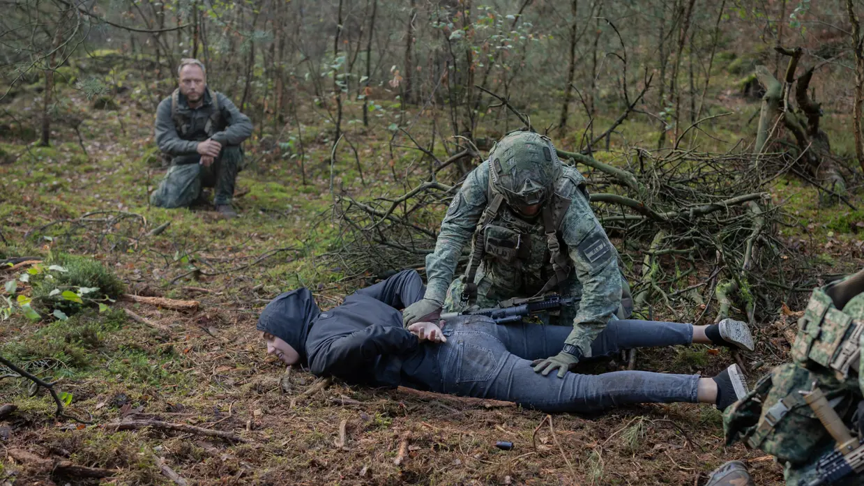 Drie gewapende militairen in gesprek met drie personen in donkere burgerkleding. Een vrouw met een donkere trui en een bedekt gezicht. Militairen in gevecht met de donker geklede partizanen. Twee militairen houden een persoon in burgerkleding vast terwijl diegene met zijn buik op de grond ligt.