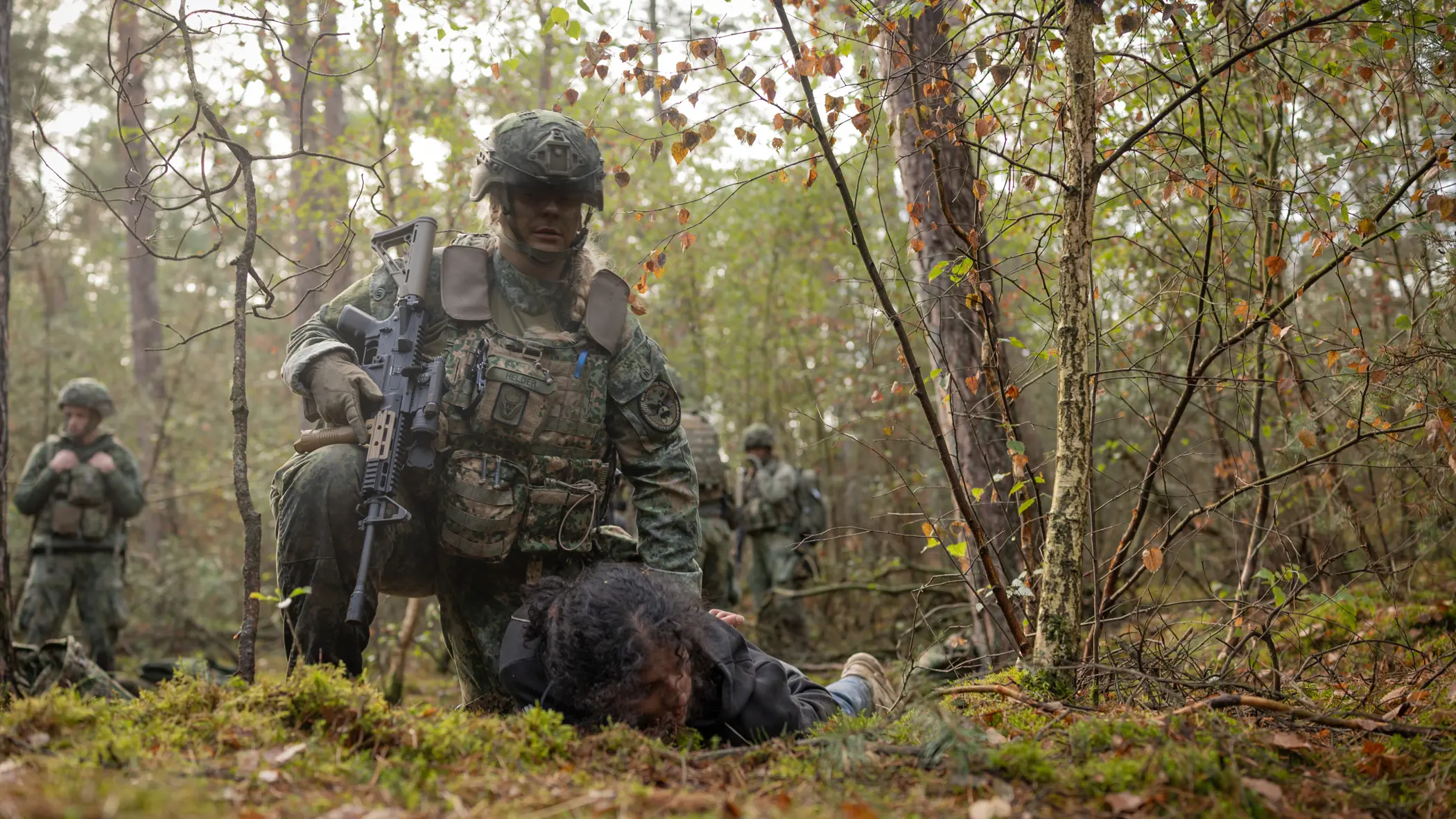 Twee militairen in het bos houden ieder een persoon in burgerkleding tegen de grond gedrukt.