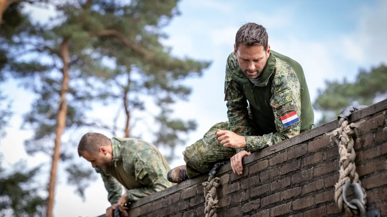 Een sportinstructeur wijst, terwijl achter hem militairen over een muurtje klimmen. Twee militairen bewegen zich over en onder een obstakel met horizontale palen door. Een militair springt over een obstakel op de hindernisbaan. Twee militairen kruipen door een obstakel op de hindernisbaan.