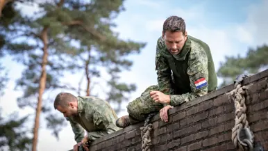 Een sportinstructeur wijst, terwijl achter hem militairen over een muurtje klimmen. Twee militairen bewegen zich over en onder een obstakel met horizontale palen door. Een militair springt over een obstakel op de hindernisbaan. Twee militairen kruipen door een obstakel op de hindernisbaan.