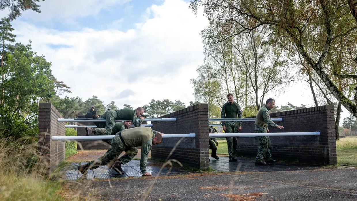 Een sportinstructeur wijst, terwijl achter hem militairen over een muurtje klimmen. Twee militairen bewegen zich over en onder een obstakel met horizontale palen door. Een militair springt over een obstakel op de hindernisbaan. Twee militairen kruipen door een obstakel op de hindernisbaan.