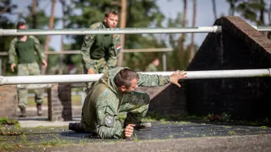 Een sportinstructeur wijst, terwijl achter hem militairen over een muurtje klimmen. Twee militairen bewegen zich over en onder een obstakel met horizontale palen door. Een militair springt over een obstakel op de hindernisbaan. Twee militairen kruipen door een obstakel op de hindernisbaan.