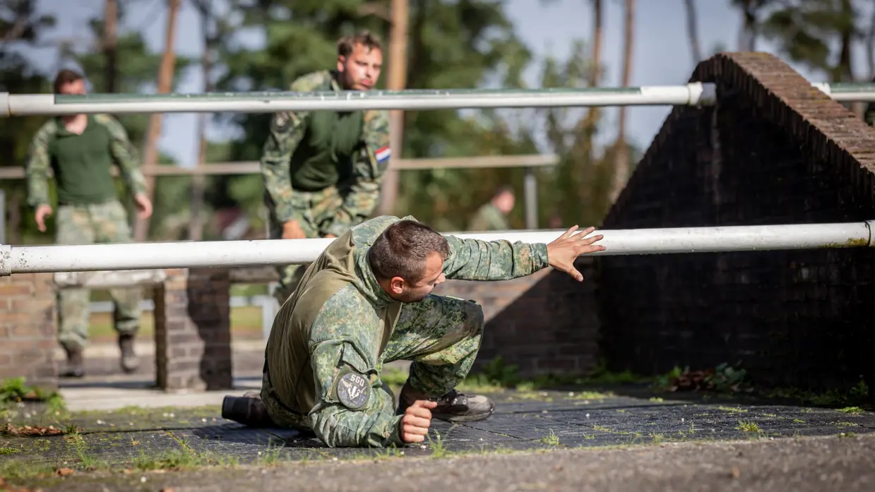 Een sportinstructeur wijst, terwijl achter hem militairen over een muurtje klimmen. Twee militairen bewegen zich over en onder een obstakel met horizontale palen door. Een militair springt over een obstakel op de hindernisbaan. Twee militairen kruipen door een obstakel op de hindernisbaan.