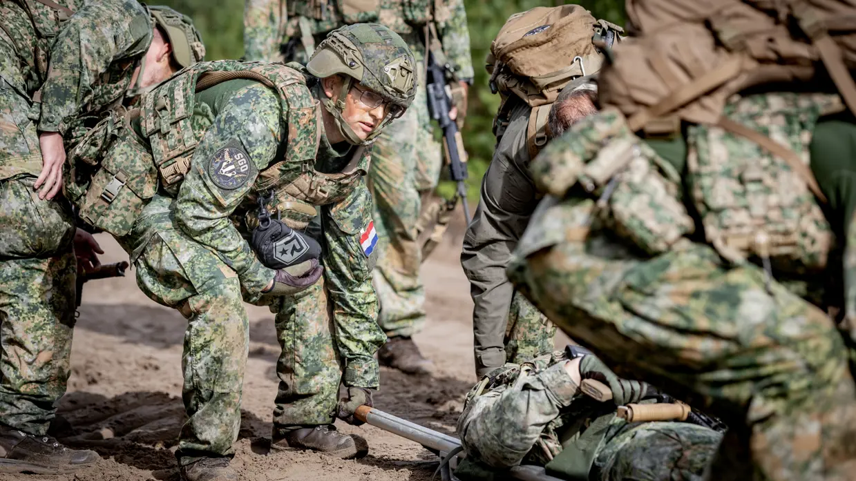 Een groepje militairen tilt een brancard van de grond op een zanderige weg. De militairen lopen met de brancard.