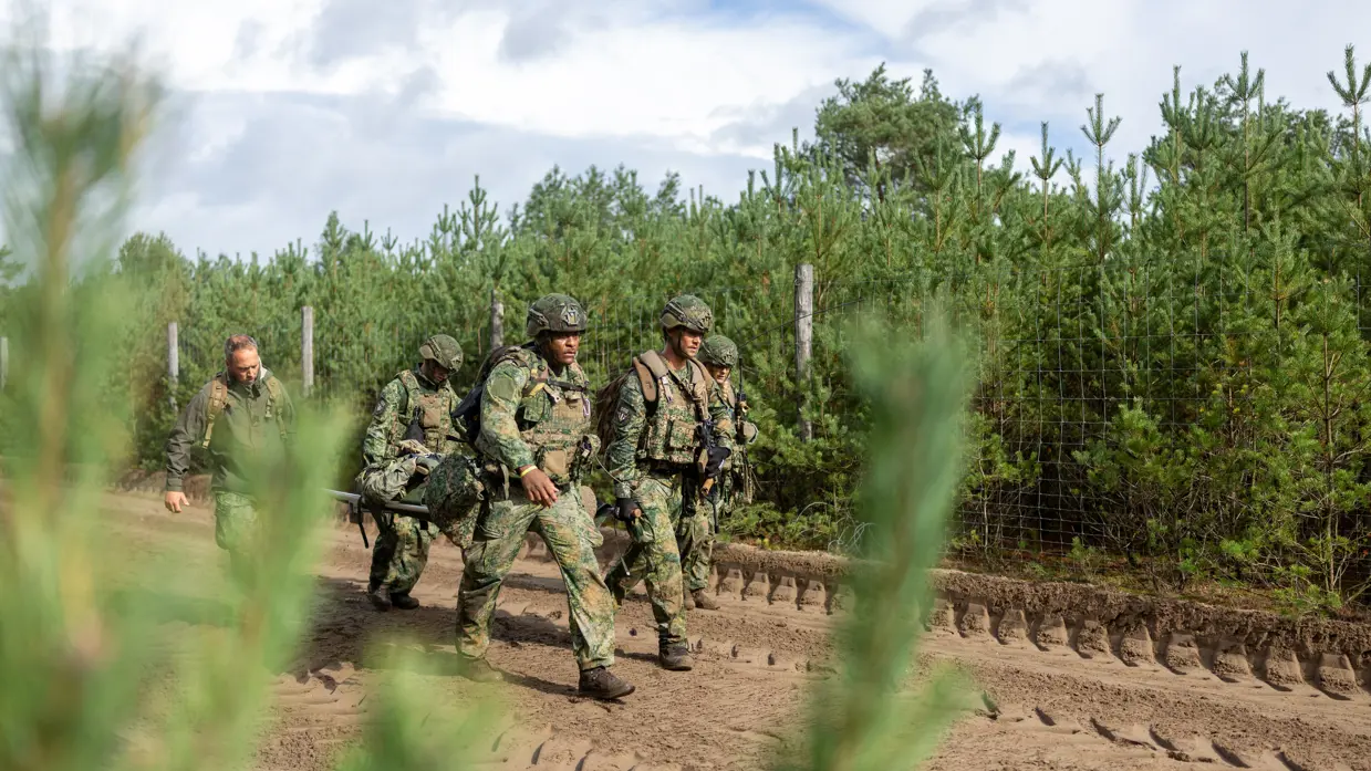 Een groepje militairen tilt een brancard van de grond op een zanderige weg. De militairen lopen met de brancard.