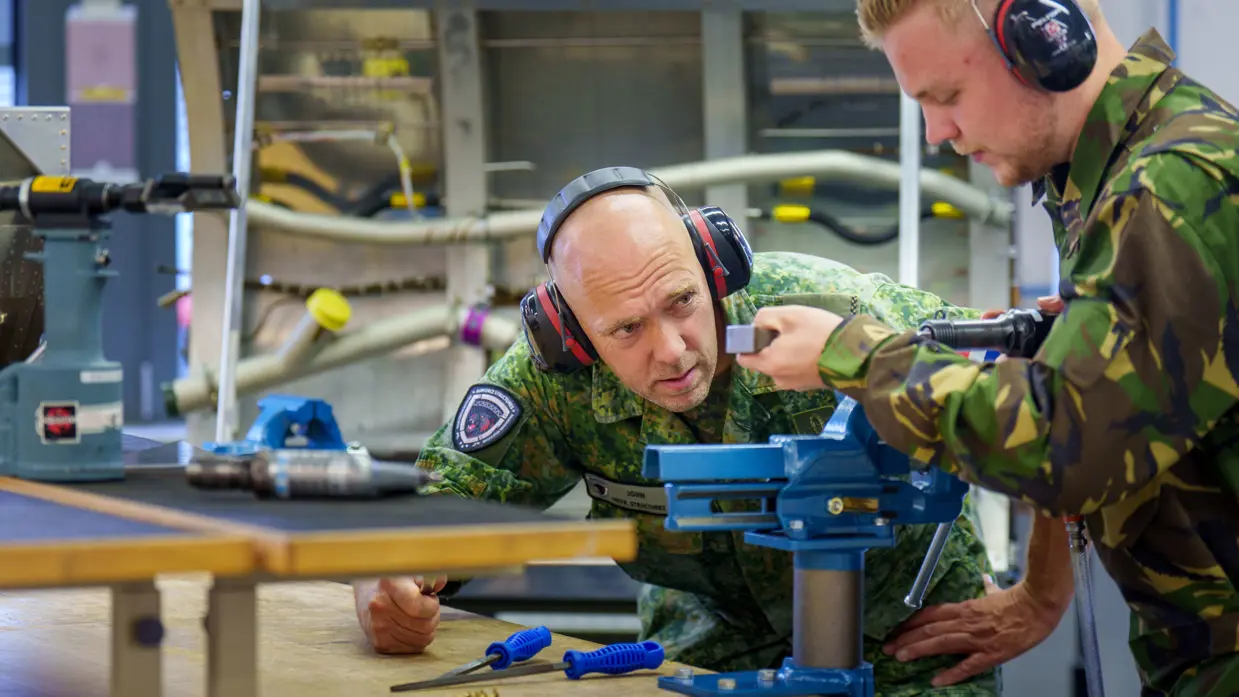 Fotocollage van John Butijn aan het werk als instructeur. Eenmaal heeft hij z’n armen over elkaar en kijkt hij in de camera. Een ander moment helpt hij een leerling bij een aambeeld en zit hij met leerlingen in een gedemonteerde helikopter.