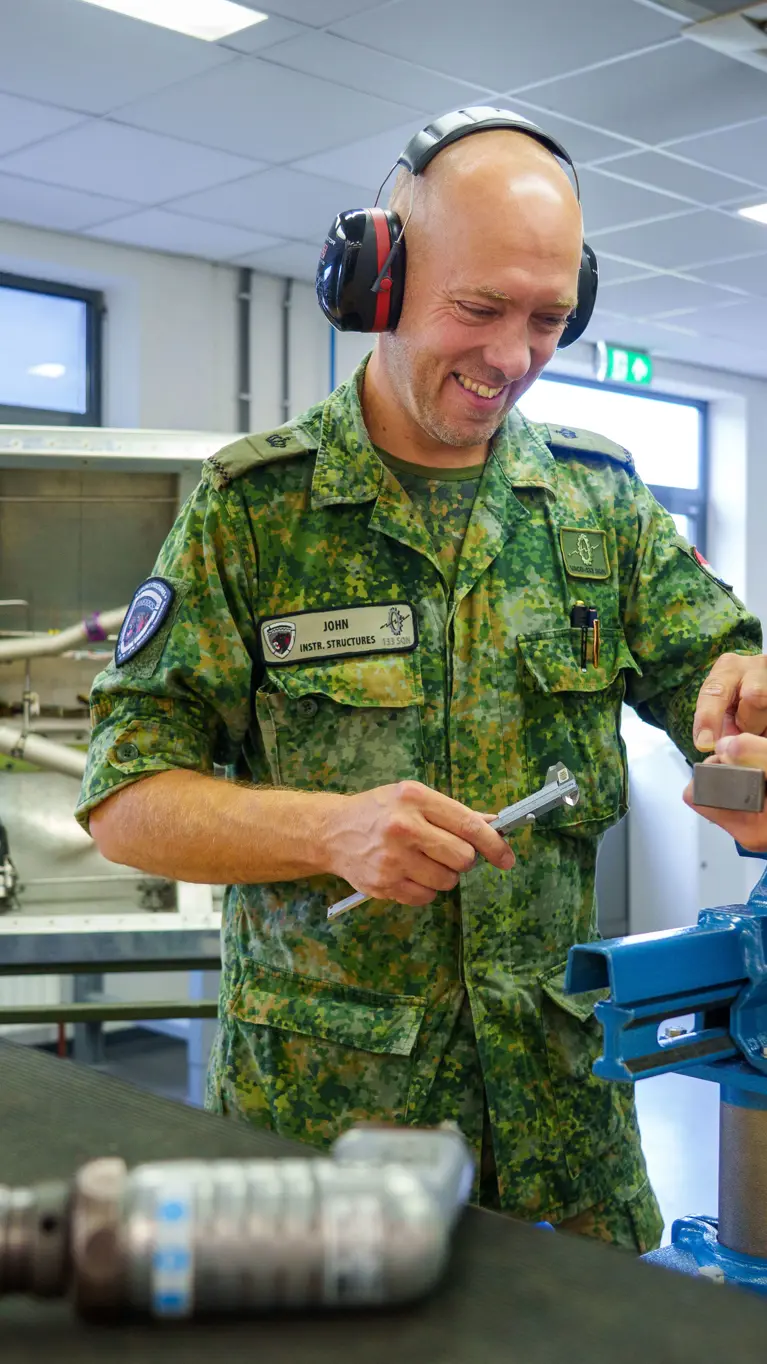 Twee militairen in pak buigen zich over een zilverkleurig stuk ijzer en dragen beide een koptelefoon. Ze staan op een werkplaats.