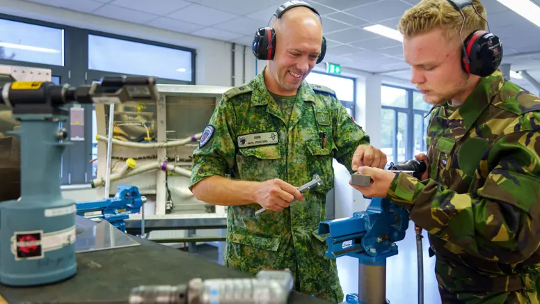 Twee militairen in pak buigen zich over een zilverkleurig stuk ijzer en dragen beide een koptelefoon. Ze staan op een werkplaats.
