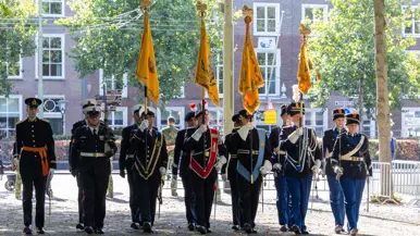  Klein groepje militairen in ceremonieel tenue. Vier van hen dragen een oranje vaandel. Ze staan in een stedelijke omgeving met bebouwing en bomen.