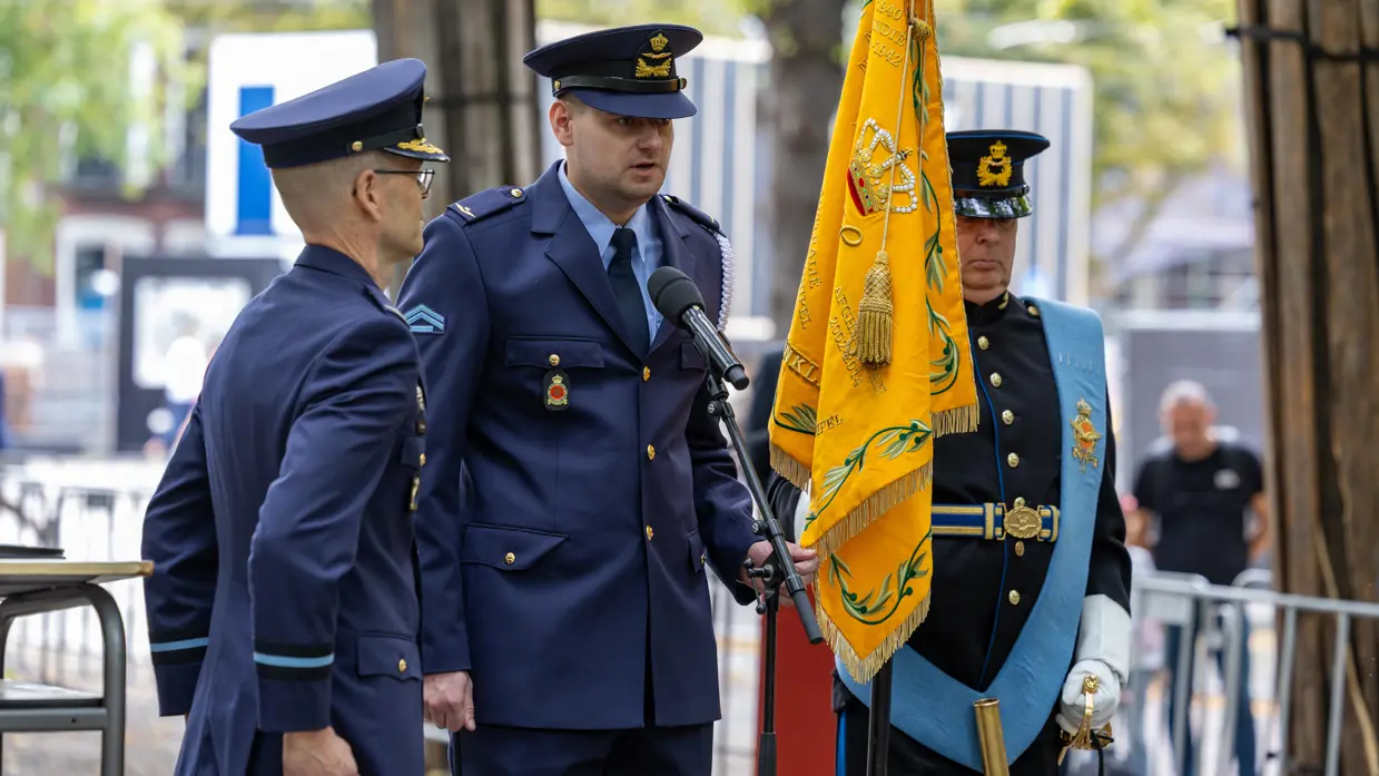  Een luchtmachtmilitair spreekt door een microfoon op standaard terwijl hij de punt van een oranje vaandel vasthoudt. Rechts staat een militair die het vaandel vasthoudt en links een luchtmachtmilitair in de houding.