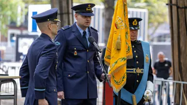  Een luchtmachtmilitair spreekt door een microfoon op standaard terwijl hij de punt van een oranje vaandel vasthoudt. Rechts staat een militair die het vaandel vasthoudt en links een luchtmachtmilitair in de houding.