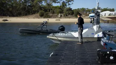Een grijze, vliegende drone onder een strakblauwe lucht. Een man bestuurt een zwart, onbemand bootje vanaf de kade. Een zwart bootje vaart weg vanaf de kade. Een oranje torpedo ligt in de golvende zee.