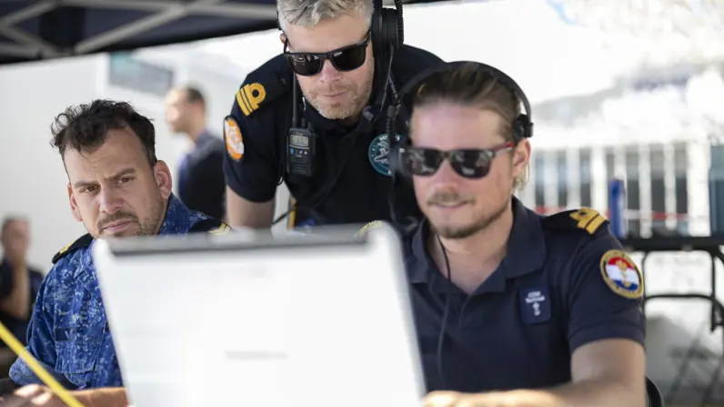 Drie militairen van de marine kijken naar een computer. In het midden een grijze man met zonnebril op.&nbsp;