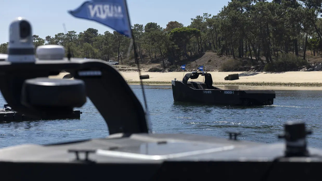Een grijze, vliegende drone onder een strakblauwe lucht. Een man bestuurt een zwart, onbemand bootje vanaf de kade. Een zwart bootje vaart weg vanaf de kade. Een oranje torpedo ligt in de golvende zee.