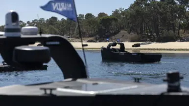 Een grijze, vliegende drone onder een strakblauwe lucht. Een man bestuurt een zwart, onbemand bootje vanaf de kade. Een zwart bootje vaart weg vanaf de kade. Een oranje torpedo ligt in de golvende zee.