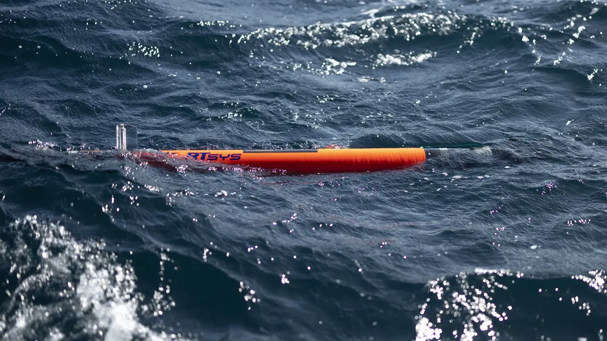 Een grijze, vliegende drone onder een strakblauwe lucht. Een man bestuurt een zwart, onbemand bootje vanaf de kade. Een zwart bootje vaart weg vanaf de kade. Een oranje torpedo ligt in de golvende zee.