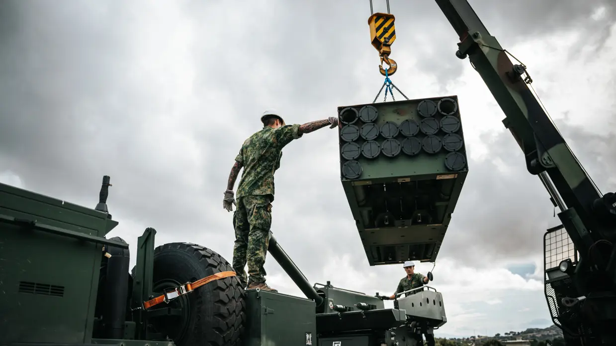 Een hijskraan tilt een soort container in de lucht, dit gebeurt onder begeleiding van twee militairen met een witte helm op.