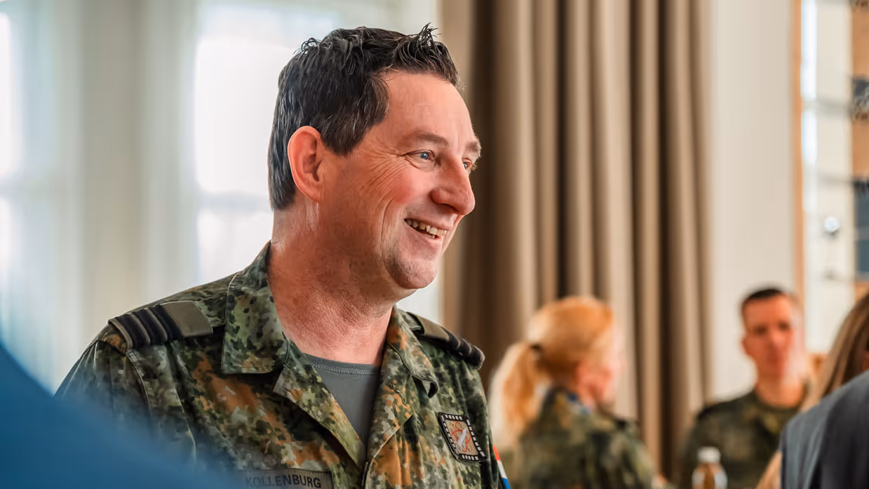 Een Nederlandse militair kijkt lachend naar rechts, op de achtergrond een paar militairen in gesprek.