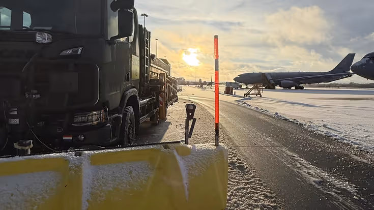 Een sneeuwschuiver ruimt een besneeuwde startbaan op luchthaven Eindhoven, terwijl op de achtergrond een vliegtuig en de ondergaande zon zichtbaar zijn.