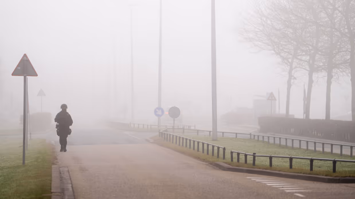 Een persoon in donkere kleding en uitrusting loopt op een mistige weg langs verkeersborden; bomen en lantaarnpalen verdwijnen in dichte, grijze mist.