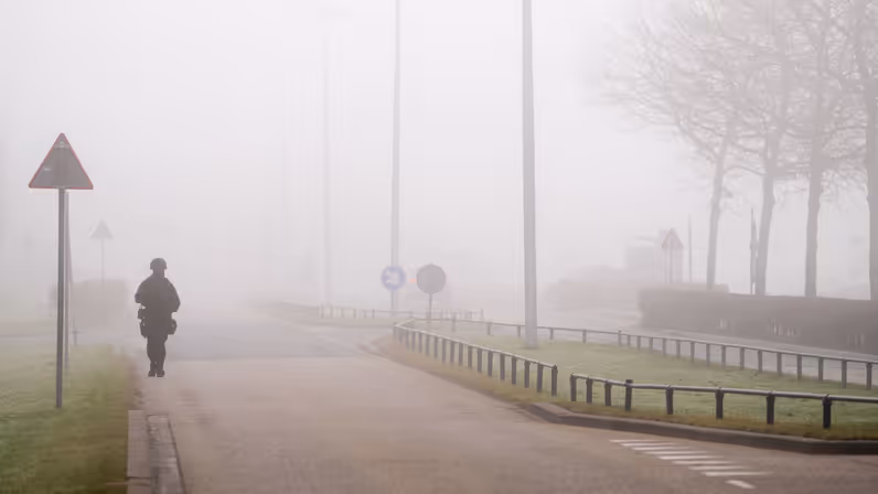 Een persoon in donkere kleding en uitrusting loopt op een mistige weg langs verkeersborden; bomen en lantaarnpalen verdwijnen in dichte, grijze mist.