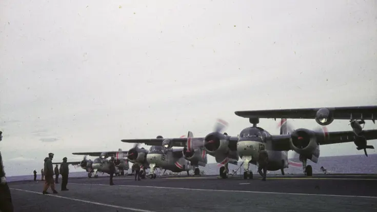Vijf vliegtuigen staan met draaiende propellers op een vliegdek, met mannen eromheen en zee op de achtergrond.